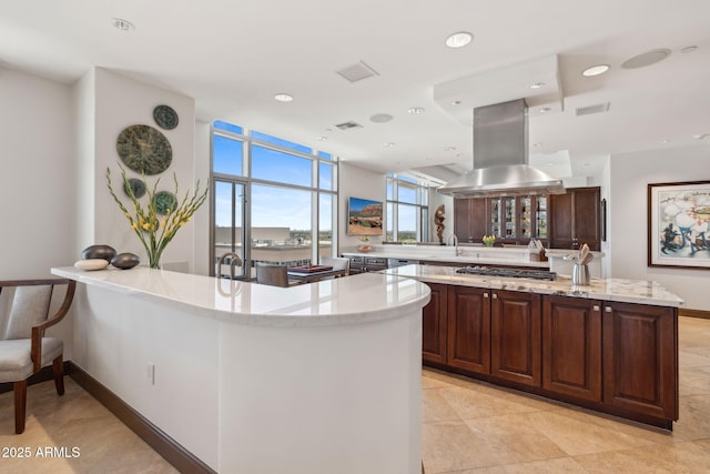 kitchen featuring island range hood, a sink, stainless steel gas cooktop, light stone countertops, and a peninsula
