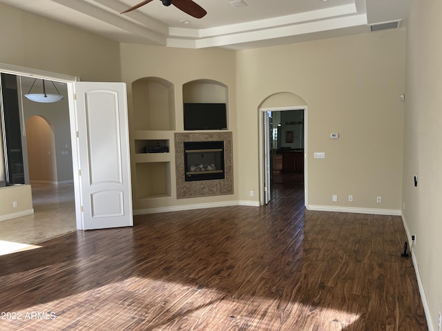unfurnished living room featuring built in shelves, dark hardwood / wood-style flooring, a tray ceiling, and ceiling fan