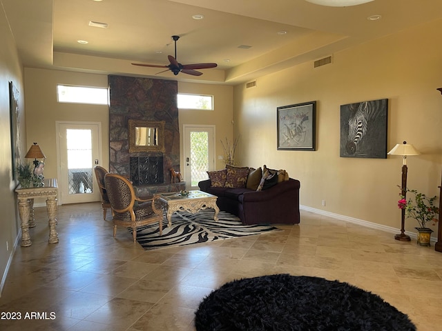 living room featuring a raised ceiling, a wealth of natural light, a stone fireplace, and ceiling fan