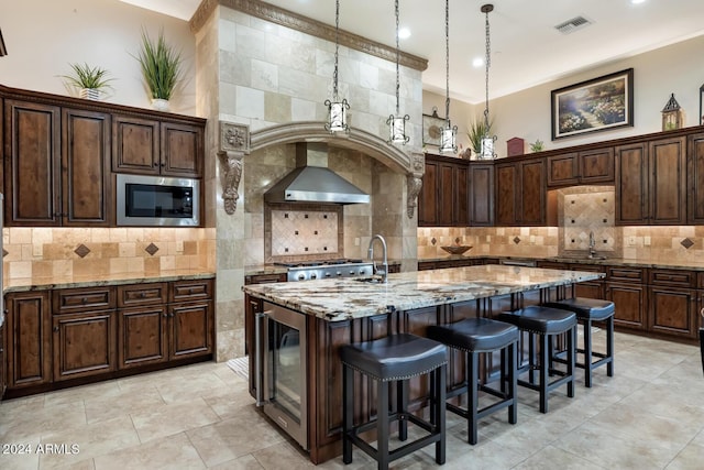 kitchen with a kitchen island with sink, beverage cooler, wall chimney exhaust hood, stainless steel microwave, and a sink
