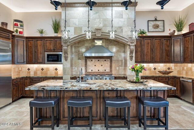 kitchen featuring decorative backsplash, built in appliances, wall chimney range hood, and light stone countertops