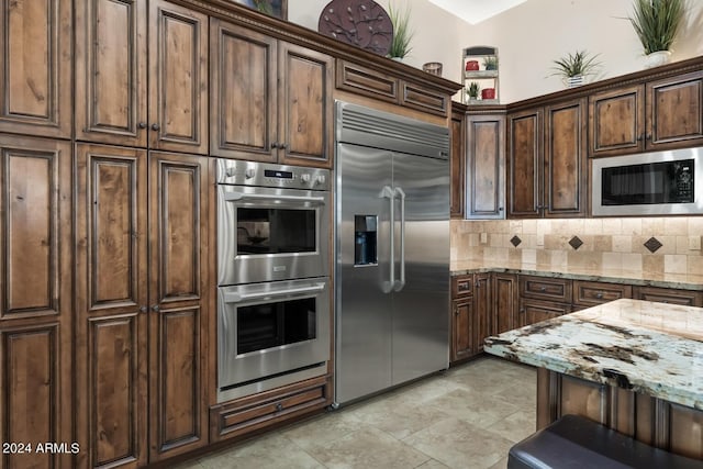 kitchen featuring built in appliances, decorative backsplash, light stone counters, and dark brown cabinets