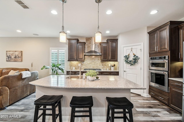 kitchen with appliances with stainless steel finishes, a kitchen island with sink, dark brown cabinets, and wall chimney range hood