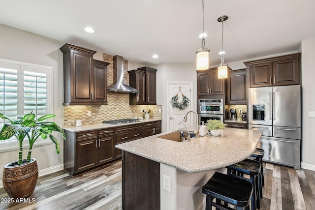 kitchen with appliances with stainless steel finishes, sink, wall chimney range hood, dark brown cabinetry, and a center island with sink