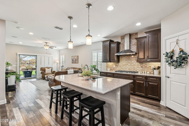 kitchen with dark brown cabinetry, a breakfast bar, stainless steel gas cooktop, a kitchen island with sink, and wall chimney range hood