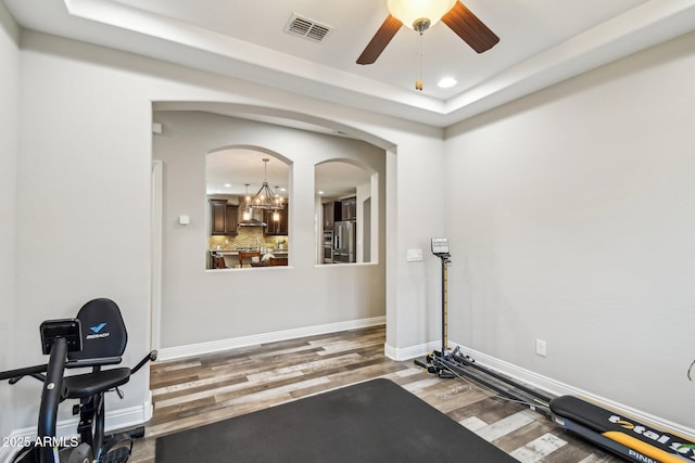 workout area with dark hardwood / wood-style flooring, ceiling fan with notable chandelier, and a raised ceiling