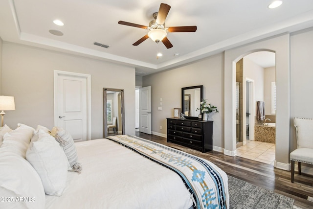 bedroom featuring ceiling fan, ensuite bath, wood-type flooring, and a tray ceiling