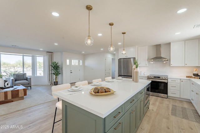 kitchen featuring stainless steel appliances, white cabinets, wall chimney range hood, a center island, and light wood finished floors