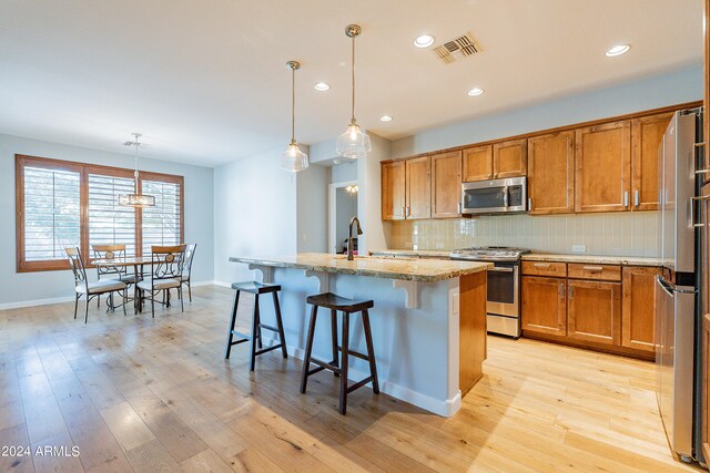 kitchen featuring light stone counters, light hardwood / wood-style floors, an island with sink, hanging light fixtures, and appliances with stainless steel finishes