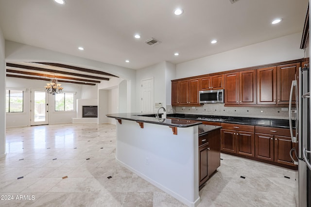 kitchen featuring beam ceiling, a kitchen island with sink, backsplash, appliances with stainless steel finishes, and a kitchen bar