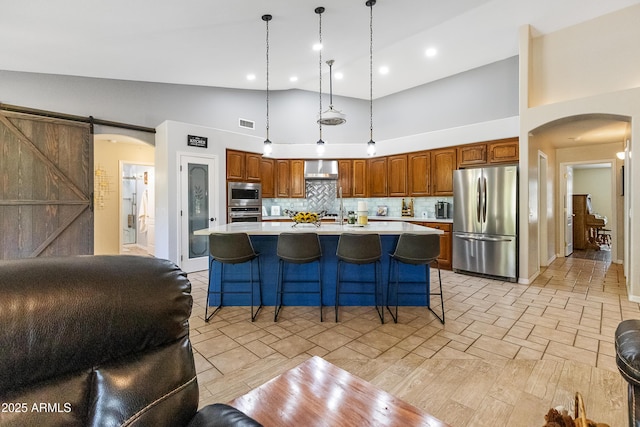 kitchen with backsplash, stainless steel appliances, a center island with sink, decorative light fixtures, and a barn door