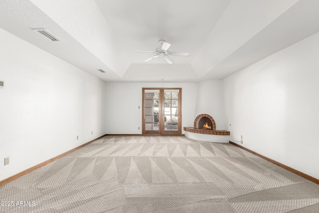 unfurnished room featuring a large fireplace, ceiling fan, a tray ceiling, light carpet, and french doors