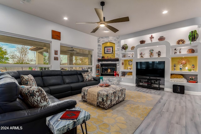 living room with built in shelves, ceiling fan, a stone fireplace, and light wood-type flooring