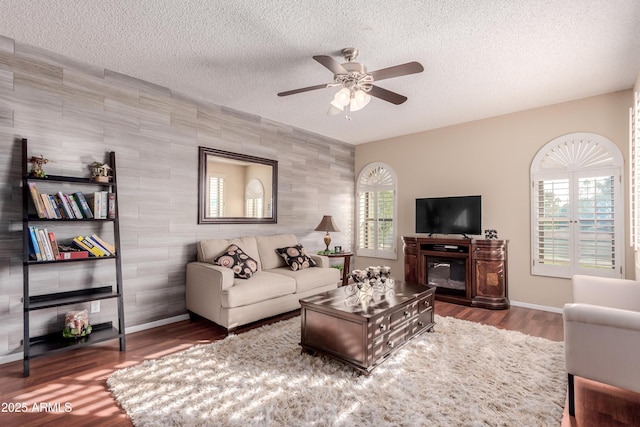 living area with a ceiling fan, a textured ceiling, a glass covered fireplace, wood finished floors, and baseboards