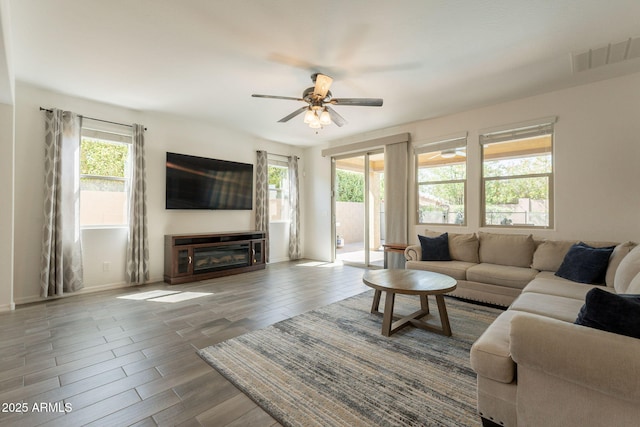 living room featuring visible vents, a glass covered fireplace, wood finished floors, baseboards, and a ceiling fan
