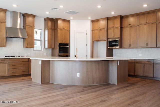 kitchen with appliances with stainless steel finishes, visible vents, wall chimney exhaust hood, brown cabinetry, and backsplash