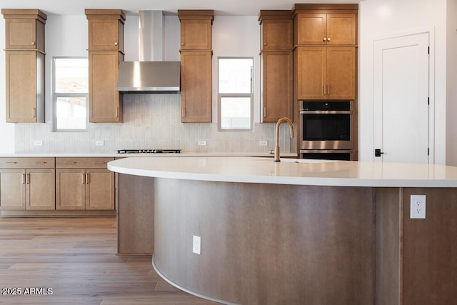 kitchen featuring wall chimney exhaust hood, light wood-style floors, double oven, and light countertops