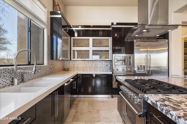 kitchen featuring tasteful backsplash, glass insert cabinets, built in appliances, island exhaust hood, and a sink