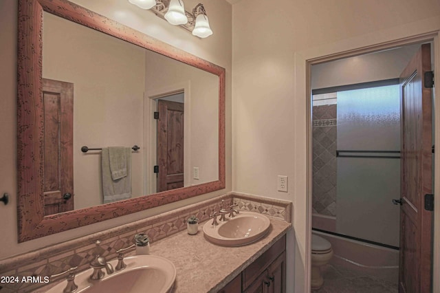 bathroom with double vanity, bath / shower combo with glass door, toilet, and a sink