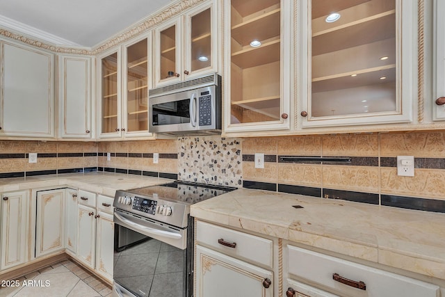 kitchen featuring light tile patterned floors, glass insert cabinets, stainless steel appliances, cream cabinetry, and light countertops