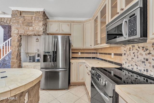 kitchen featuring glass insert cabinets, ornamental molding, cream cabinets, stainless steel appliances, and light countertops