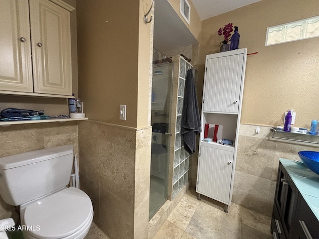 full bath featuring tiled shower, toilet, a wainscoted wall, vanity, and tile walls