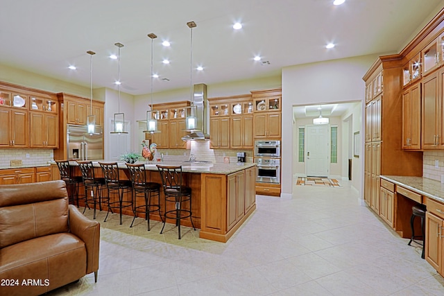 kitchen with a breakfast bar area, a large island with sink, stainless steel appliances, wall chimney range hood, and open floor plan