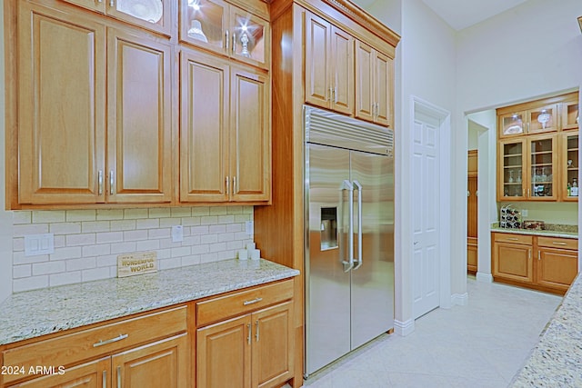 kitchen with light stone counters, light tile patterned floors, stainless steel built in fridge, glass insert cabinets, and tasteful backsplash