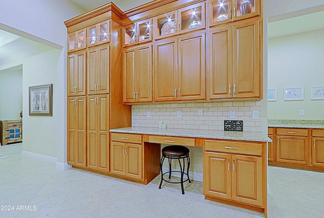 kitchen featuring light stone counters, backsplash, light tile patterned floors, glass insert cabinets, and built in study area