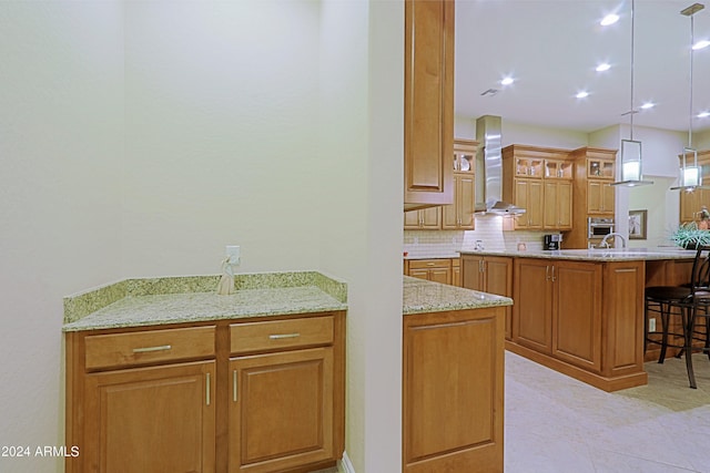 kitchen featuring brown cabinetry, light stone countertops, a kitchen bar, wall chimney range hood, and tasteful backsplash