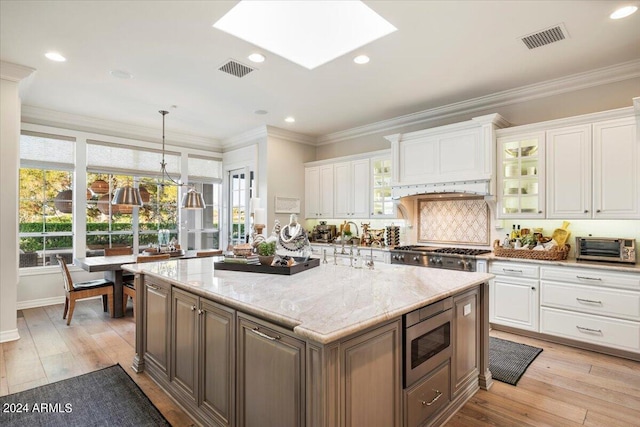 kitchen with light wood-type flooring, stainless steel microwave, a kitchen island with sink, white cabinetry, and hanging light fixtures