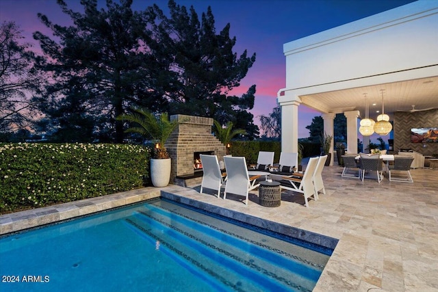 pool at dusk featuring a patio and an outdoor brick fireplace