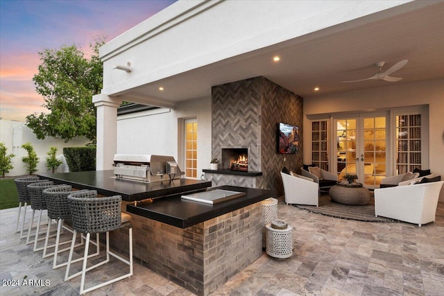 patio terrace at dusk featuring ceiling fan, an outdoor living space with a fireplace, grilling area, area for grilling, and a bar