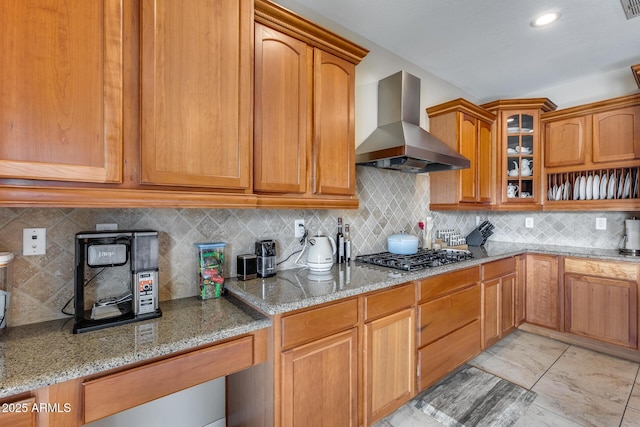 kitchen featuring backsplash, range hood, stainless steel gas stovetop, glass insert cabinets, and light stone countertops