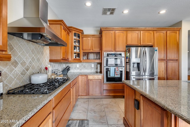 kitchen with light stone counters, visible vents, stainless steel appliances, glass insert cabinets, and wall chimney exhaust hood