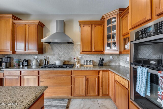 kitchen featuring light stone counters, stainless steel appliances, glass insert cabinets, and wall chimney exhaust hood