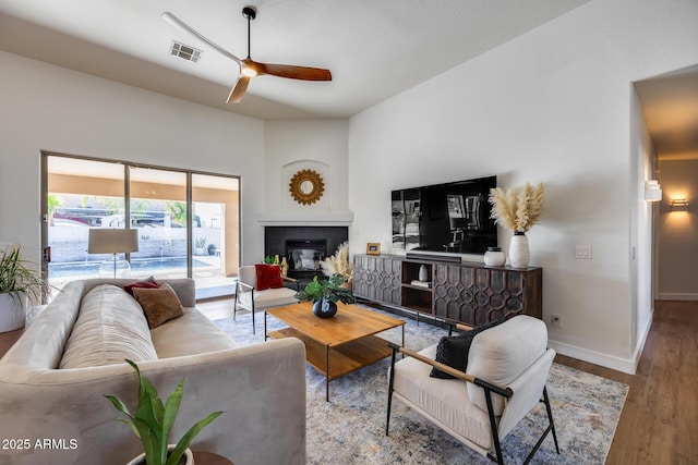 living area featuring a ceiling fan, wood finished floors, visible vents, baseboards, and a fireplace