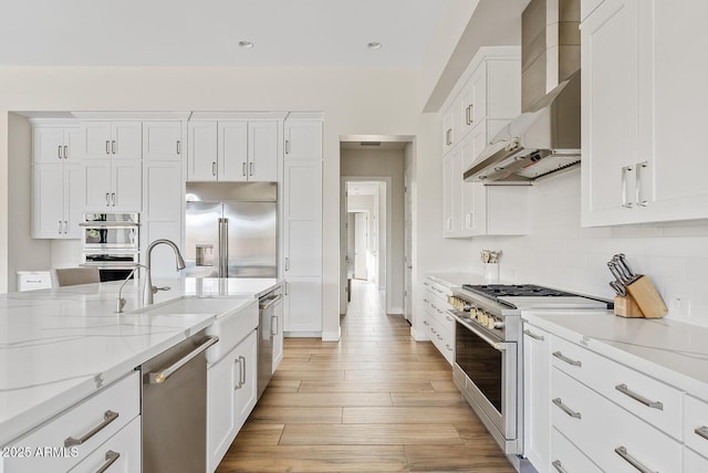 kitchen with wall chimney range hood, high quality appliances, light stone countertops, light wood finished floors, and white cabinetry