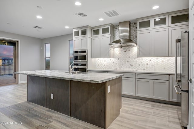 kitchen with a kitchen island with sink, wall chimney range hood, light hardwood / wood-style flooring, light stone countertops, and stainless steel appliances