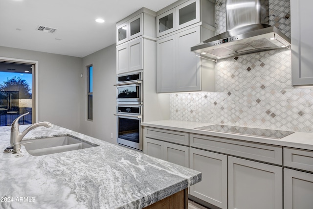 kitchen with black electric stovetop, light stone counters, wall chimney exhaust hood, stainless steel double oven, and sink