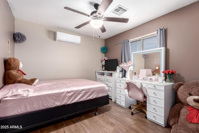 bedroom with a wall unit AC, wood finished floors, visible vents, and a ceiling fan