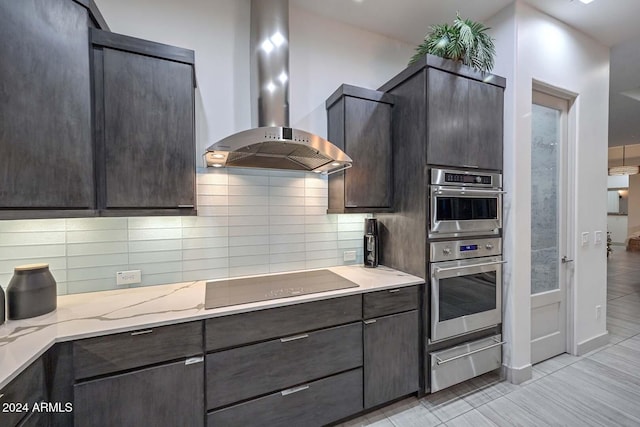 kitchen featuring wall chimney range hood, black electric cooktop, dark brown cabinets, light stone counters, and stainless steel double oven