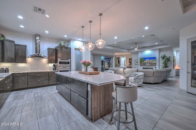 kitchen featuring tasteful backsplash, wall chimney exhaust hood, a tray ceiling, ceiling fan, and a large island