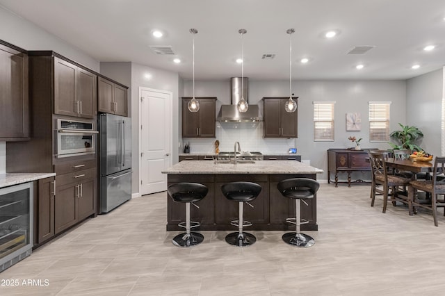 kitchen featuring appliances with stainless steel finishes, light stone countertops, an island with sink, beverage cooler, and wall chimney exhaust hood