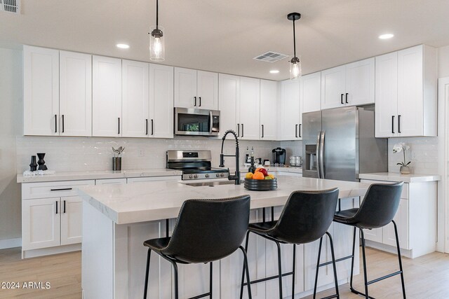 kitchen with white cabinets, a center island with sink, decorative light fixtures, stainless steel appliances, and light hardwood / wood-style floors