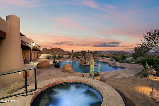 pool at dusk featuring a mountain view, a patio area, and an in ground hot tub