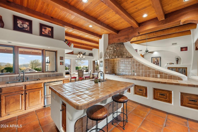 kitchen featuring wood ceiling, ceiling fan with notable chandelier, a kitchen island with sink, tile counters, and a breakfast bar area