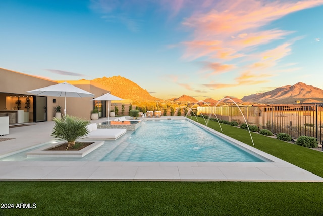 pool at dusk featuring a patio area, a mountain view, pool water feature, and a lawn