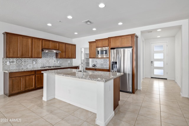 kitchen with an island with sink, stainless steel appliances, backsplash, light stone counters, and sink