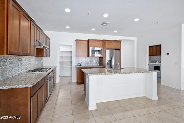 kitchen with washing machine and dryer, a kitchen island with sink, range hood, appliances with stainless steel finishes, and light stone counters